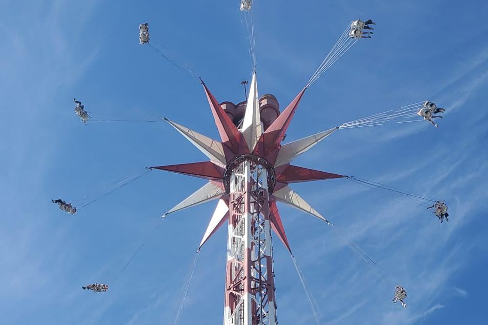 Ein hohes Kettenkarussell ragt vor einem strahlend blauen Himmel in die Höhe. Die Fahrgäste schwingen in ihren Sitzen an langen Ketten kreisförmig um den rot-weiß gestalteten Turm. Die Aufnahme wurde aus der Untersicht gemacht und betont die Höhe der Attraktion